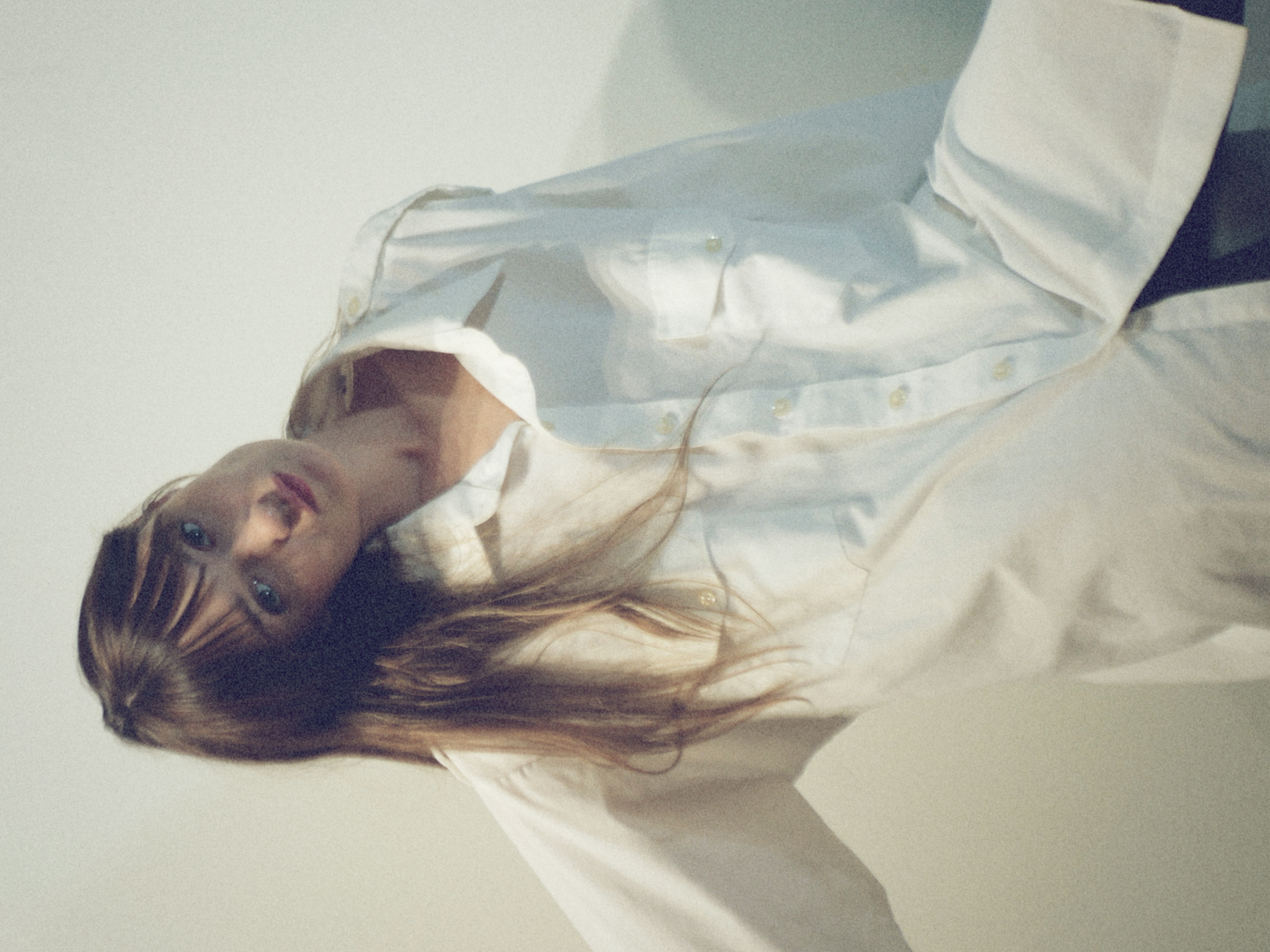 Kris Brouillette in an oversized white shirt, head tilted, one arm reaching out against a pale wall